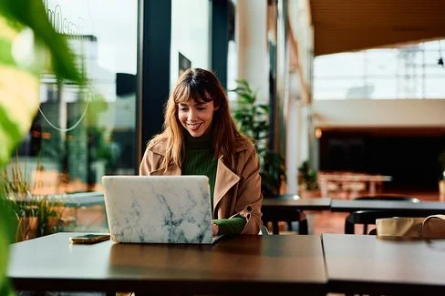 Eine Frau mit langem Haar und Mantel sitzt an einem Tisch in einem Café und arbeitet an einem Laptop.