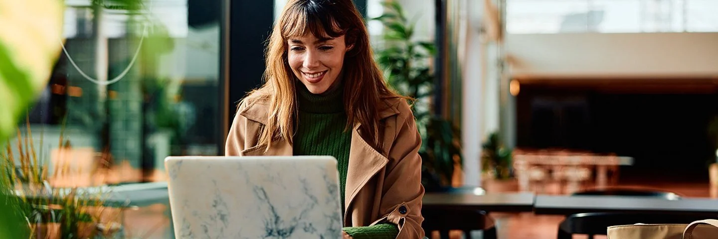 Eine Frau mit langem Haar und Mantel sitzt an einem Tisch in einem Café und arbeitet an einem Laptop.