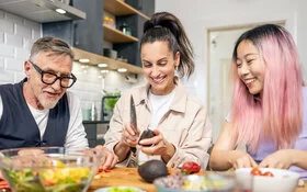 3 Personen unterschiedlichen Alters, Geschlecht und Ethnie sitzen an einem Tisch und bereiten einen Salat zu