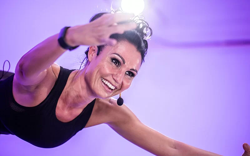 Pilates Coach Nicole Willberg ist vor lilanem Hintergrund in Nahaufnahme zu sehen und lächelt.