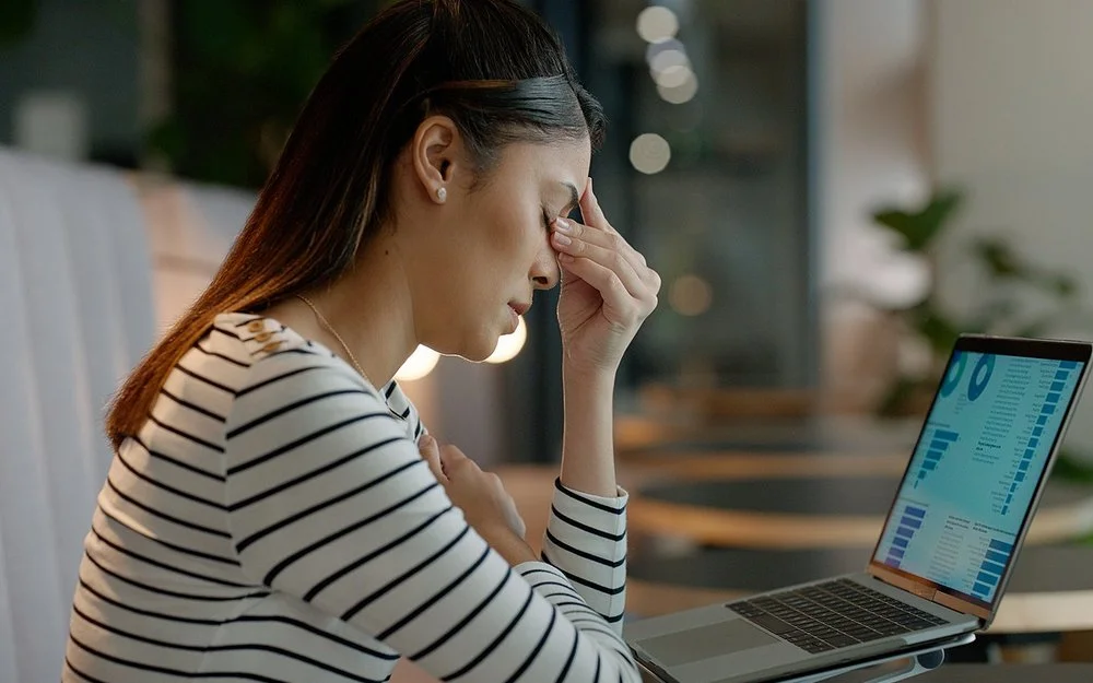 Eine Frau sitzt an einem Schreibtisch und hält sich die linke Hand an die geschlossenen Augen.