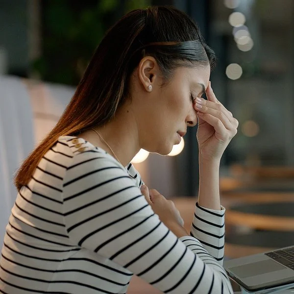 Eine Frau sitzt an einem Schreibtisch und hält sich die linke Hand an die geschlossenen Augen.