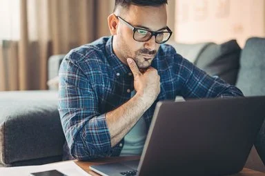 Ein junger Mann mit Brille stüzt das mit der rechten Hand ab und blickt auf den Bildschirm eines Laptops.