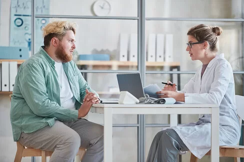 Ein blondierter Mann sitzt am Tisch bei einer Ärztin, die mit ihm spricht.