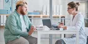 Ein blondierter Mann sitzt an einem Tisch mit einer Ärztin und lässt sich beraten.