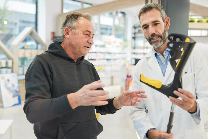 Ein Mann hebt artikulierend die Hände, während ein anderer Mann im weißen Kittel eine Gehhilfe präsentiert.