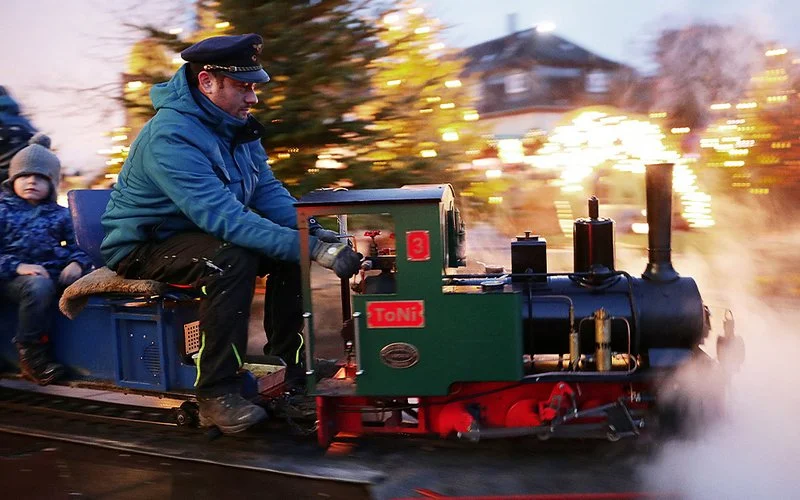 Kleine Dampflok mit Fahrgästen fährt durch den beleuchteten Weihnachtsmarkt in Bad Homburg; Dampf steigt auf.