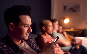 Ein Mann sitzt mit Freunden auf der Couch und checkt sein Smartphone aus Angst, etwas zu verpassen.