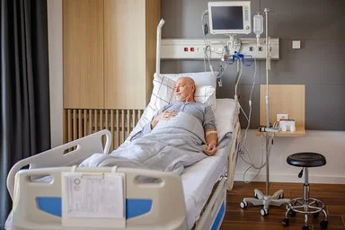 Ein älterer Mann liegt in einem Krankenhausbett in einem Patientenzimmer. Er ist an medizinische Geräte und eine Infusion angeschlossen. Er ist wach und blickt Richtung Fenster.