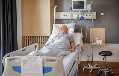 Ein älterer Mann liegt in einem Krankenhausbett in einem Patientenzimmer. Er ist an medizinische Geräte und eine Infusion angeschlossen. Er ist wach und blickt Richtung Fenster.