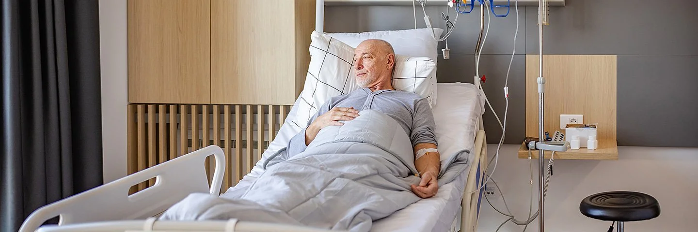 Ein älterer Mann liegt in einem Krankenhausbett in einem Patientenzimmer. Er ist an medizinische Geräte und eine Infusion angeschlossen. Er ist wach und blickt Richtung Fenster.
