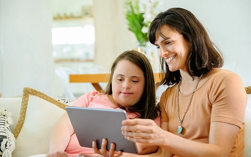 Eine Mutter und ihre Tochter mit Trisomie 21 sitzen im Wohnzimmer auf einem Sofa und schauen gemeinsam auf ein Tablet.