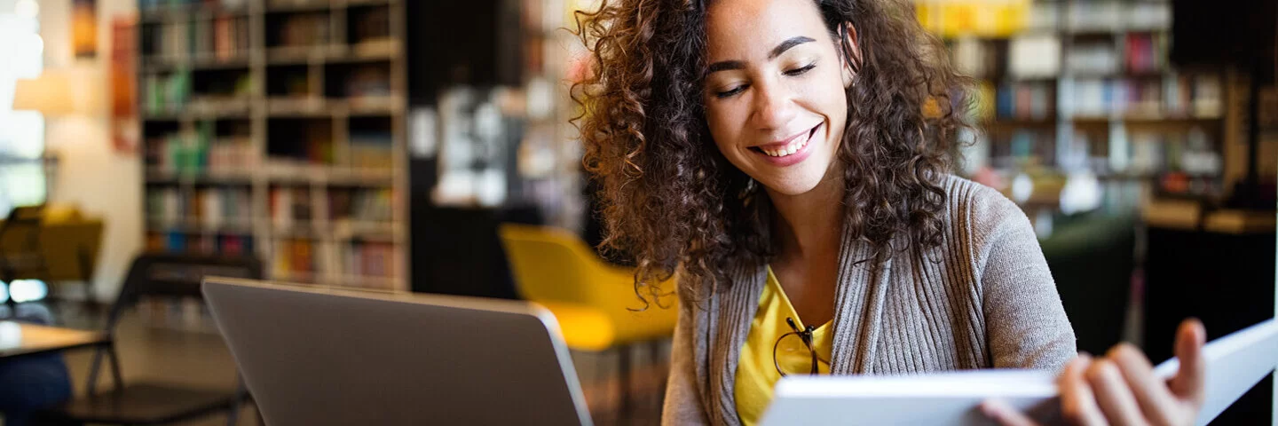 Eine junge Frau, die Stress im Studium hat, sitzt in der Bibliothek vor ihrem Laptop und ihren Lernunterlagen.