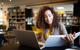 Eine junge Frau, die Stress im Studium hat, sitzt in der Bibliothek vor ihrem Laptop und ihren Lernunterlagen.
