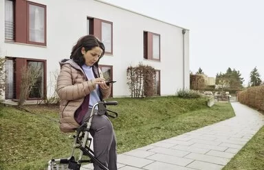 Frau mit Gehhilfe vor Haus schaut skeptisch auf Smartphone