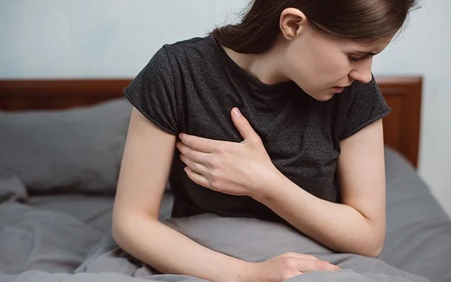 Eine Frau sitzt mit einem T-Shirt bekleidet im Bett und fasst sich seitlich an die schmerzende Brust.