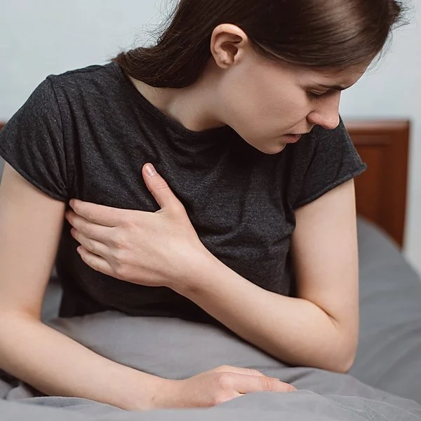 Eine Frau sitzt mit einem T-Shirt bekleidet im Bett und fasst sich seitlich an die schmerzende Brust.