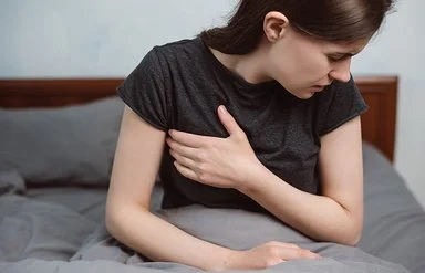 Eine Frau sitzt mit einem T-Shirt bekleidet im Bett und fasst sich seitlich an die schmerzende Brust.