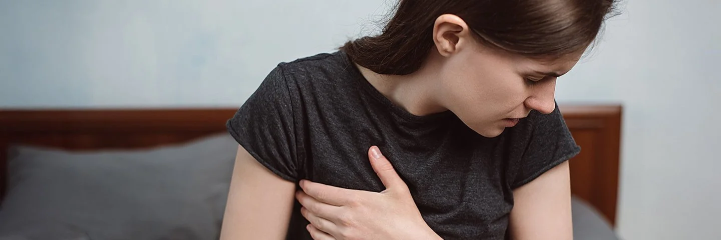 Eine Frau sitzt mit einem T-Shirt bekleidet im Bett und fasst sich seitlich an die schmerzende Brust.