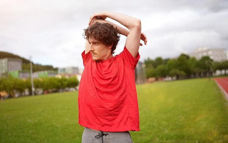 Ein junger Mann steht auf einer Wiese und verschränkt die Arme hinter dem Rücken. Die Aktion wirkt wie eine Dehnübung.