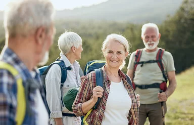 Zwei ältere Frauen und zwei ebenfalls ältere Männer sind wandern. Sie stehen an einem grünen Hang, tragen leichte Wanderrucksäcke auf den Schultern und lächeln.
