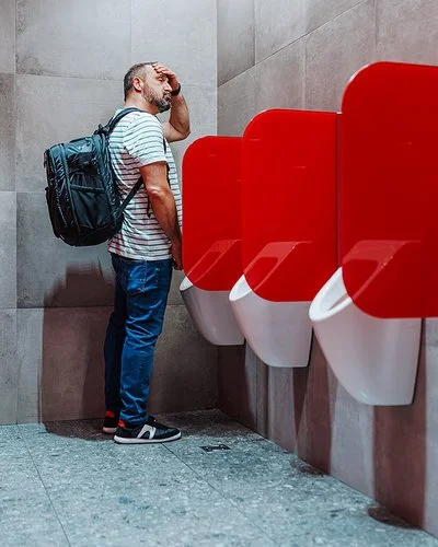 Ein mittelalter Mann steht an einem öffentlichen Urinal und fasst sich an die Stirn.