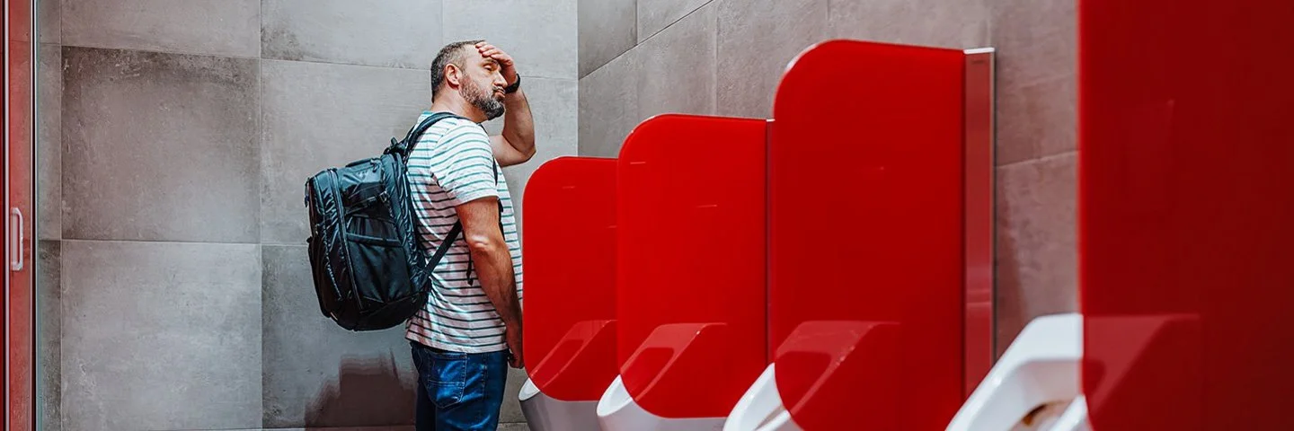 Ein mittelalter Mann steht an einem öffentlichen Urinal und fasst sich an die Stirn.