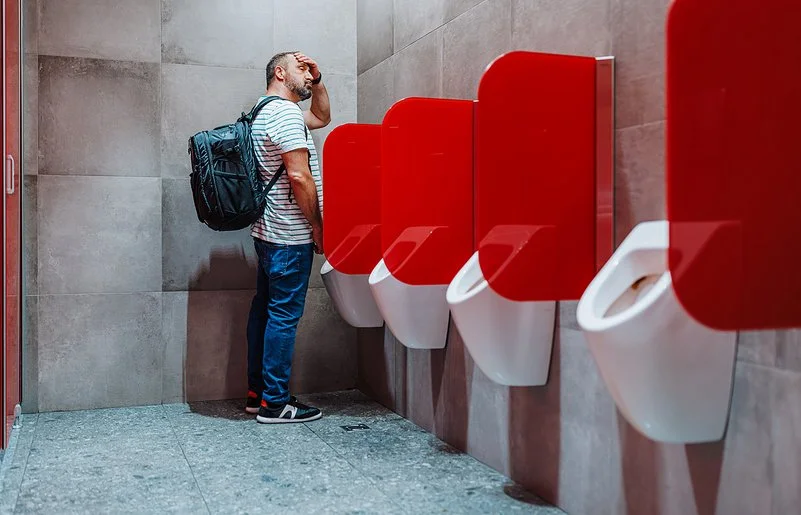 Ein mittelalter Mann steht an einem öffentlichen Urinal und fasst sich an die Stirn.