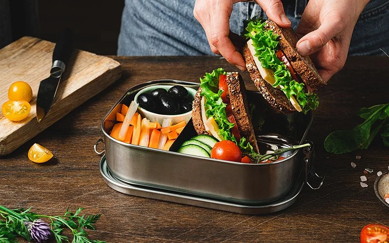 Eine Person füllt eine Brotdose mit einem Butterbrot und frischem Gemüse.