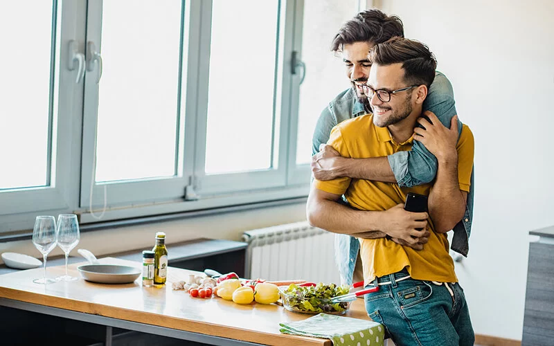 Zwei Männer kochen gemeinsam und umarmen sich dabei glücklich verliebt.