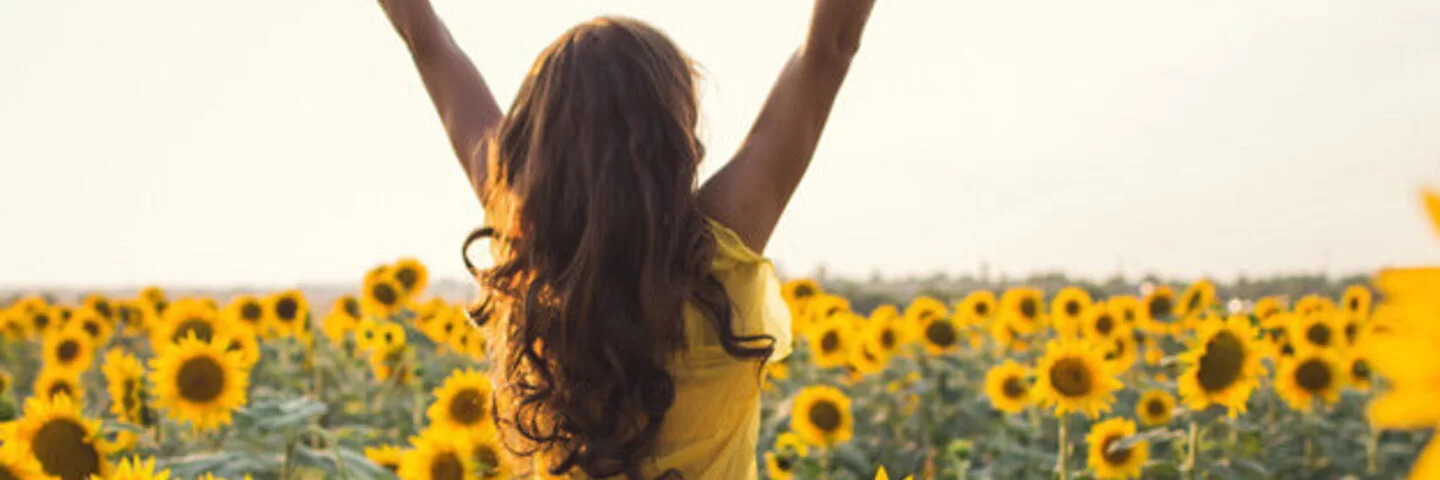 Zu sehen ist eine junge Frau die ihre Arme in einem Sonnenblumenfeld in die Höhe streckt.