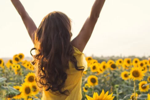 Zu sehen ist eine junge Frau die ihre Arme in einem Sonnenblumenfeld in die Höhe streckt.