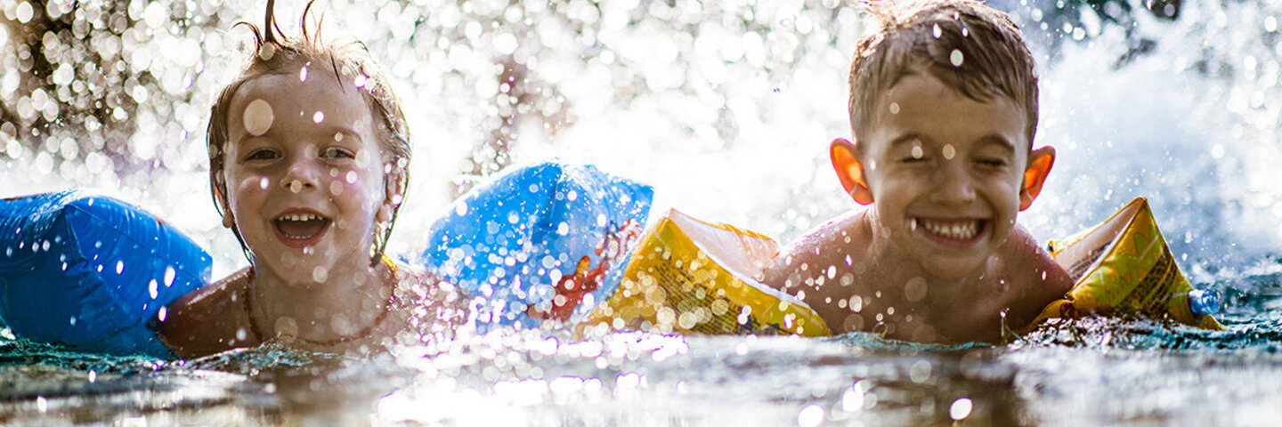 Zwei Kinder planschen im Wasser und tragen Schwimmflügel.