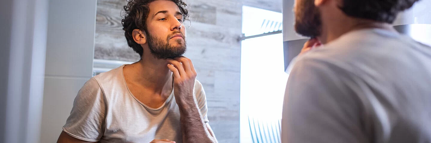 Ein junger Mann steht vor dem Badezimmerspiegel und begutachtet seinen Bart.