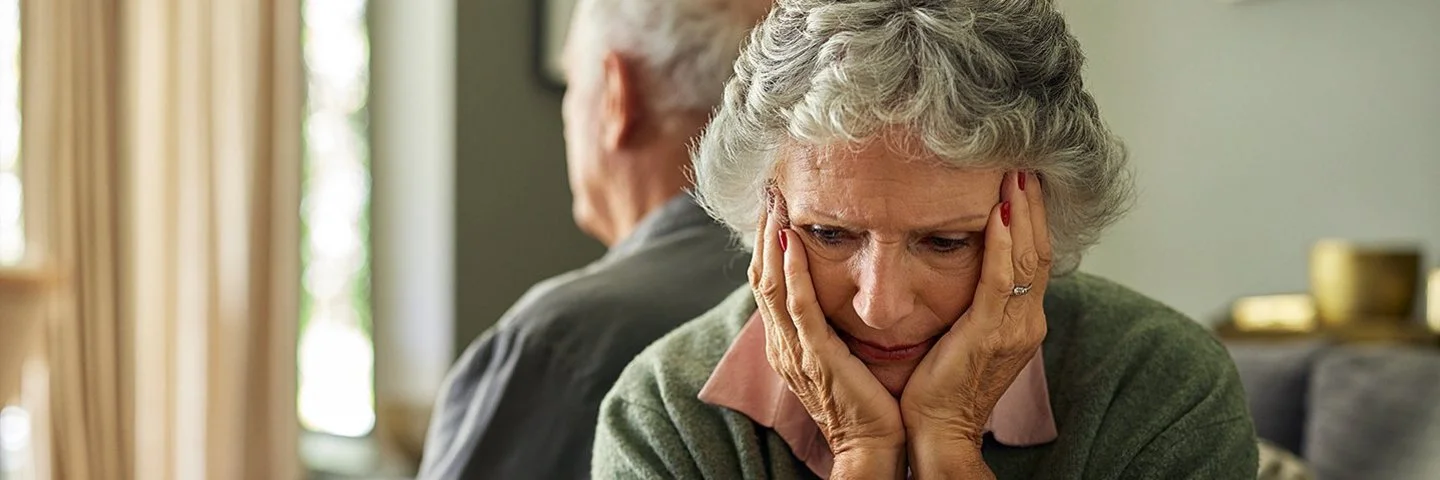 Eine ältere Frau und ihr Partner sitzen auf dem Sofa. Mit dem Rücken haben sich voneinander abgewendet. Sie hält den Kopf in ihren Händen.