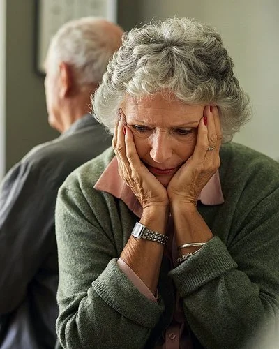Eine ältere Frau und ihr Partner sitzen auf dem Sofa. Mit dem Rücken haben sich voneinander abgewendet. Sie hält den Kopf in ihren Händen.