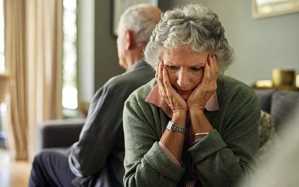 Eine ältere Frau und ihr Partner sitzen auf dem Sofa. Mit dem Rücken haben sich voneinander abgewendet. Sie hält den Kopf in ihren Händen.