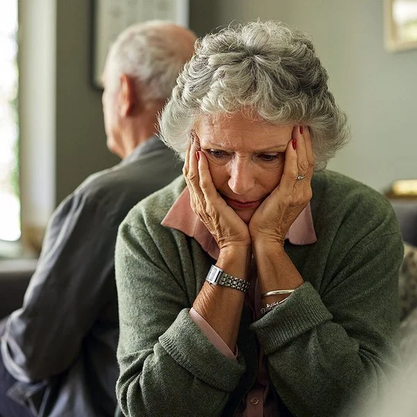Eine ältere Frau und ihr Partner sitzen auf dem Sofa. Mit dem Rücken haben sich voneinander abgewendet. Sie hält den Kopf in ihren Händen.