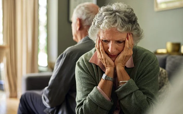 Eine ältere Frau und ihr Partner sitzen auf dem Sofa. Mit dem Rücken haben sich voneinander abgewendet. Sie hält den Kopf in ihren Händen.