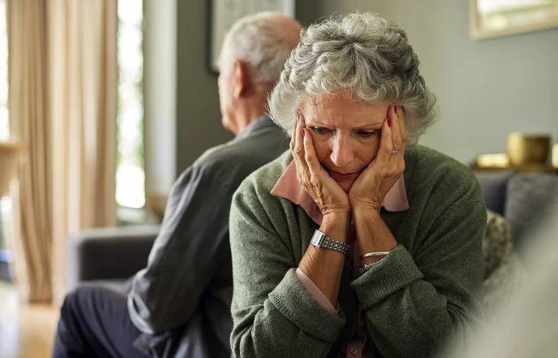 Eine ältere Frau und ihr Partner sitzen auf dem Sofa. Mit dem Rücken haben sich voneinander abgewendet. Sie hält den Kopf in ihren Händen.