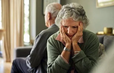 Eine ältere Frau und ihr Partner sitzen auf dem Sofa. Mit dem Rücken haben sich voneinander abgewendet. Sie hält den Kopf in ihren Händen.