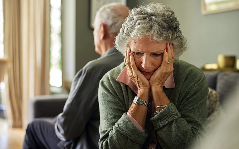 Eine ältere Frau und ihr Partner sitzen auf dem Sofa. Mit dem Rücken haben sich voneinander abgewendet. Sie hält den Kopf in ihren Händen.