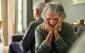 Eine ältere Frau und ihr Partner sitzen auf dem Sofa. Mit dem Rücken haben sich voneinander abgewendet. Sie hält den Kopf in ihren Händen.