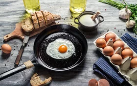 Ein rustikaler Holztisch von oben; darauf steht eine gusseiserne Pfanne mit einem frisch gebratenen Spiegelei, daneben aufgeschnittenes Brot und Eier im Karton.