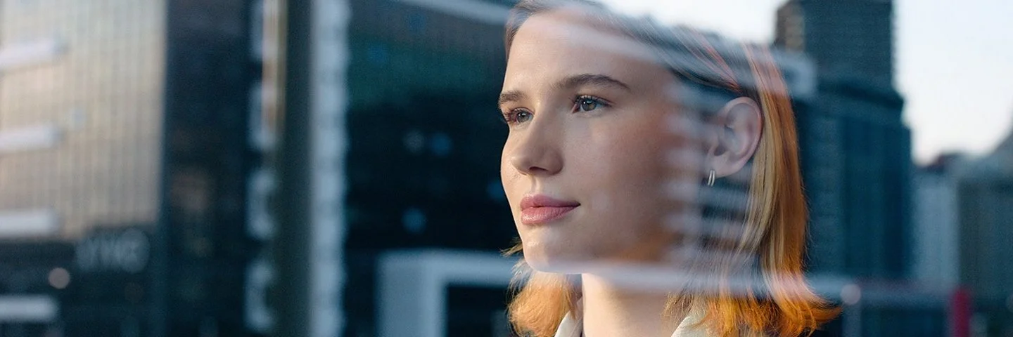 Eine junge Frau in Blazer und weißer Bluse schaut durch eine Fensterscheibe, in der sich ein Bürogebäude spiegelt, in die Ferne.