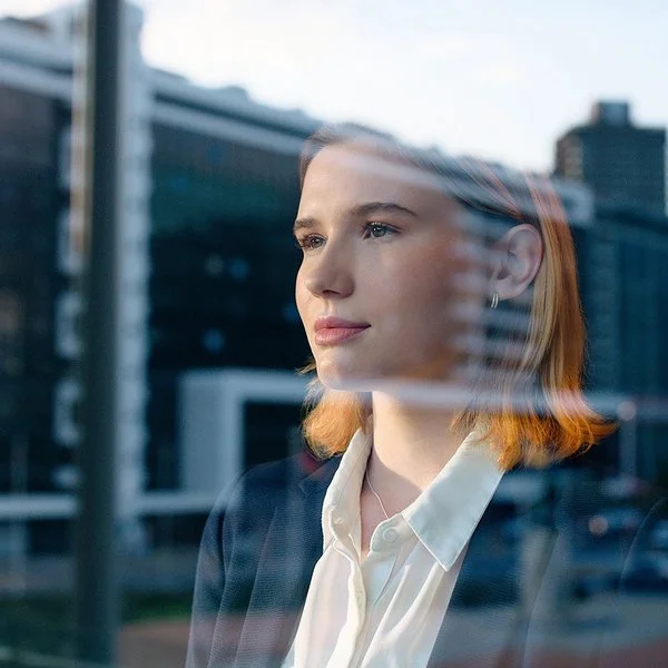 Eine junge Frau in Blazer und weißer Bluse schaut durch eine Fensterscheibe, in der sich ein Bürogebäude spiegelt, in die Ferne.