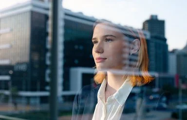 Eine junge Frau in Blazer und weißer Bluse schaut durch eine Fensterscheibe, in der sich ein Bürogebäude spiegelt, in die Ferne.