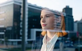 Eine junge Frau in Blazer und weißer Bluse schaut durch eine Fensterscheibe, in der sich ein Bürogebäude spiegelt, in die Ferne.