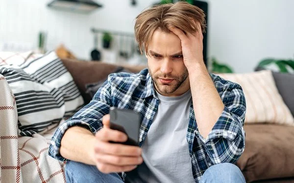 Ein junger Mann sitzt auf dem Fußboden vor einem Sofa. In der rechten Hand hält er ein Smartphone, mit der linken fährt er sich durch die Haare. Er schaut ernst auf das Display.