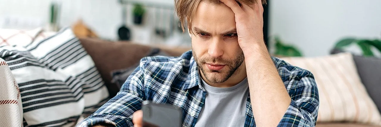 Ein junger Mann sitzt auf dem Fußboden vor einem Sofa. In der rechten Hand hält er ein Smartphone, mit der linken fährt er sich durch die Haare. Er schaut ernst auf das Display.